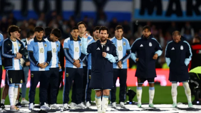 Lionel Messi berjaket timnas Argentina mengangkat tangan di lapangan dikelilingi rekan setim