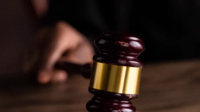 Tangan memegang palu kayu bercincin emas di atas meja dengan latar gelap