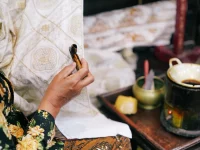 Tangan perajin memegang canting di depan kain batik dan tungku malam