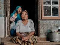 Ilustrasi perempuan lanjut usia duduk bersila di ambang pintu rumah berdinding bambu, Bandung