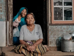 Ilustrasi perempuan lanjut usia duduk bersila di ambang pintu rumah berdinding bambu, Bandung