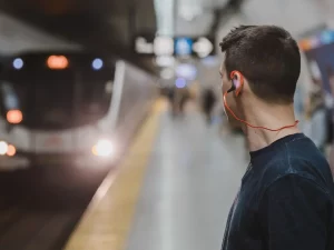 Ilustrasi pria memakai earphone berkabel merah berdiri di peron saat kereta datang
