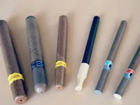 Ilustrasi enam rokok elektrik berbagai bentuk berjajar di atas meja kayu terang