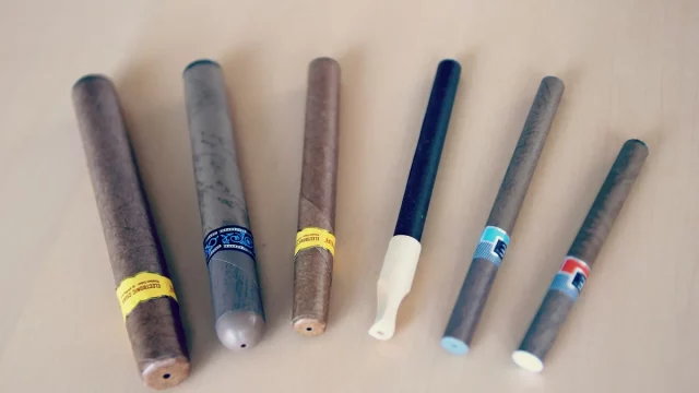 Ilustrasi enam rokok elektrik berbagai bentuk berjajar di atas meja kayu terang