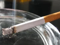 Ilustrasi sebatang rokok menyala bersandar di tepi asbak kaca dengan abu dan asap tipis