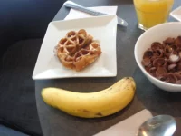 Ilustrasi menu sarapan berupa sereal cokelat, wafel, pisang, dan segelas jus jeruk di meja
