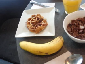 Ilustrasi menu sarapan berupa sereal cokelat, wafel, pisang, dan segelas jus jeruk di meja
