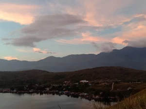 Ilustrasi kampung di tepi Danau Sentani saat senja berlatar pegunungan, Papua