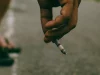 Ilustrasi tangan seorang pria menjepit rokok menyala di atas jalan aspal di Madiun