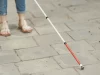 Ilustrasi kaki seseorang bersandal dengan tongkat putih berujung merah menyentuh trotoar berubin