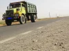 Ilustrasi truk jungkit kuning-hijau melaju di jalan raya dua lajur berlatar padang kering