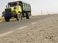 Ilustrasi truk jungkit kuning-hijau melaju di jalan raya dua lajur berlatar padang kering