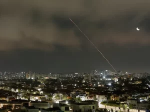 Jejak rudal melintasi langit malam di atas kota, terlihat cahaya ledakan di kejauhan selama serangan Iran-Israel