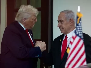 Donald Trump dan Benjamin Netanyahu berjabat tangan di Gedung Putih dengan bendera Amerika Serikat dan Israel di latar belakang