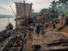 Ilustrasi pelabuhan sungai di delta Brantas dengan kapal dagang berlayar segi empat bersandar, kuli memikul bal kain bercelup nila menuruni titian papan, gudang beratap daun dan teras bata merah di tepi muara